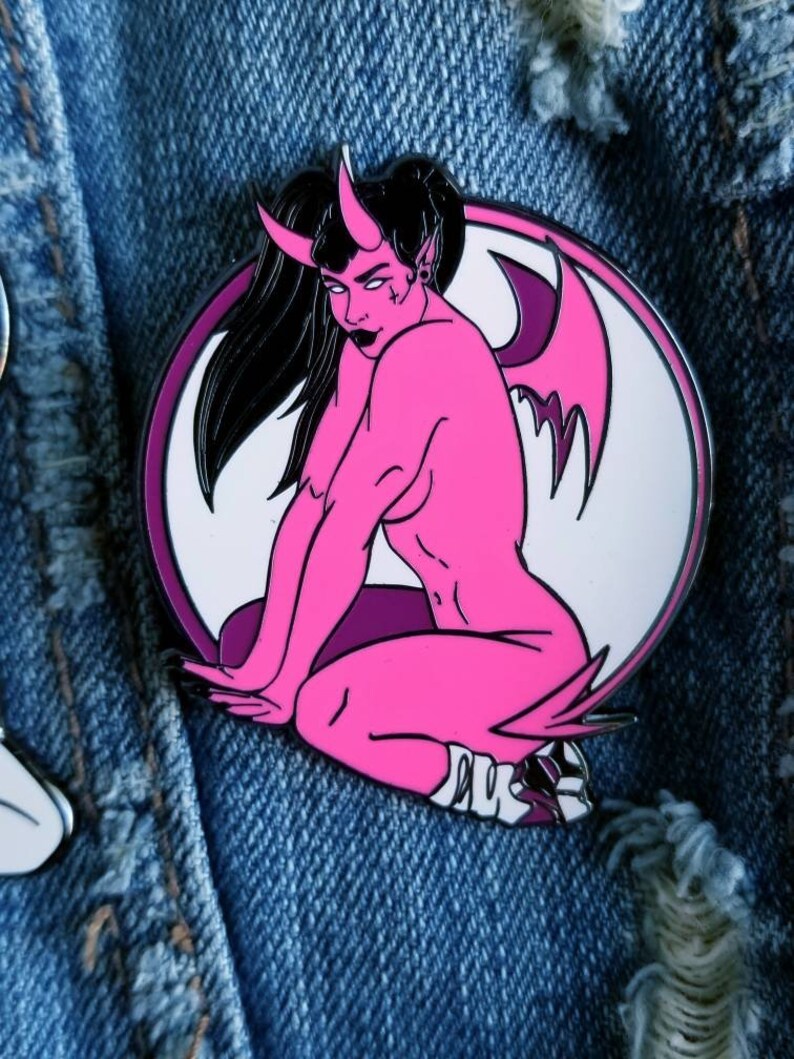 Puede incluir: Pin de esmalte rosa de una mujer demonio con alas, sentada con las piernas cruzadas. El pin est&aacute; sobre un fondo de mezclilla.