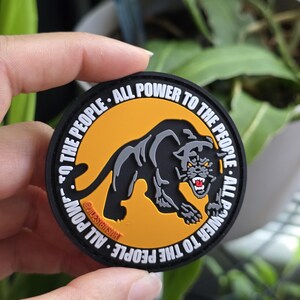 Puede incluir: Un parche redondo con una ilustraci&oacute;n de una pantera negra sobre un fondo naranja. El texto "ALL POWER TO THE PEOPLE" en blanco rodea la imagen. El parche est&aacute; sujeto por una mano.