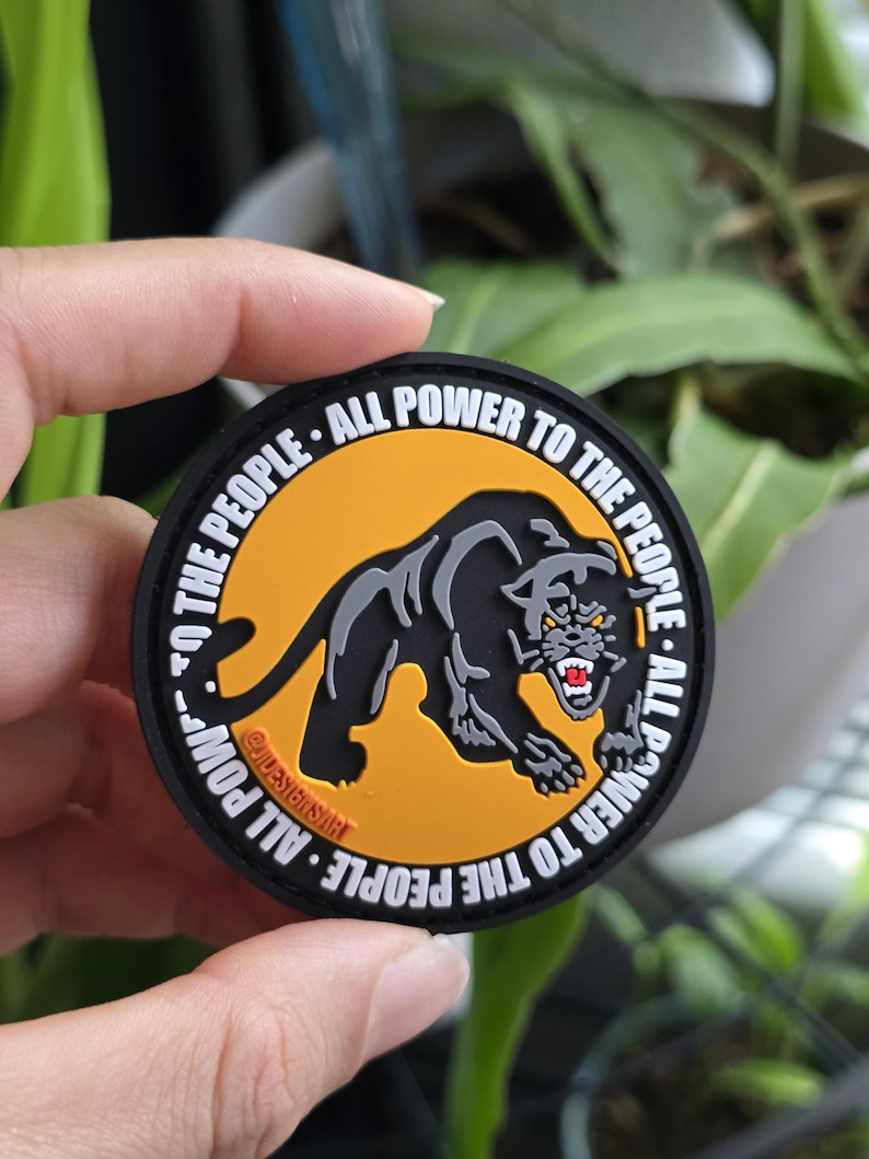 Puede incluir: Un parche redondo con una ilustraci&oacute;n de una pantera negra sobre un fondo naranja. El texto "ALL POWER TO THE PEOPLE" en blanco rodea la imagen. El parche est&aacute; sujeto por una mano.