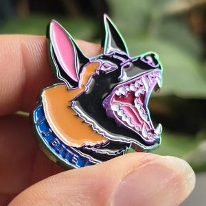 May include: A detailed enamel pin featuring a snarling dog head with sharp teeth. The pin has a black, orange, and pink color scheme with iridescent accents. The blue collar reads "I BITE".
