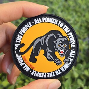 Puede incluir: Un parche redondo con una ilustraci&oacute;n de pantera negra sobre un fondo amarillo. El texto "ALL POWER TO THE PEOPLE" est&aacute; impreso en blanco alrededor del borde. El parche tiene un borde negro y tambi&eacute;n se ve el texto "@DESIGNSART".