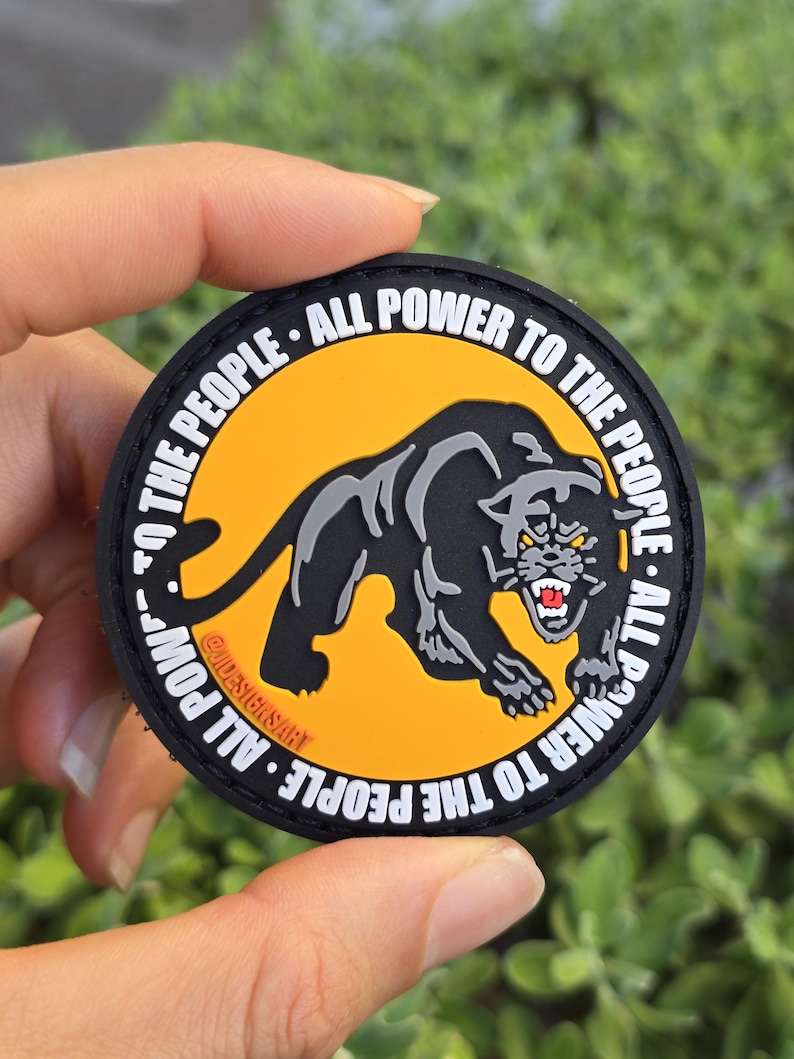 Puede incluir: Un parche redondo con una ilustraci&oacute;n de pantera negra sobre un fondo amarillo. El texto "ALL POWER TO THE PEOPLE" est&aacute; impreso en blanco alrededor del borde. El parche tiene un borde negro y tambi&eacute;n se ve el texto "@DESIGNSART".