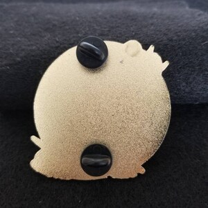 Puede incluir: La parte posterior de un pin de metal dorado con dos respaldos de goma negra. El pin presenta un dise&ntilde;o detallado de un personaje de dibujos animados.