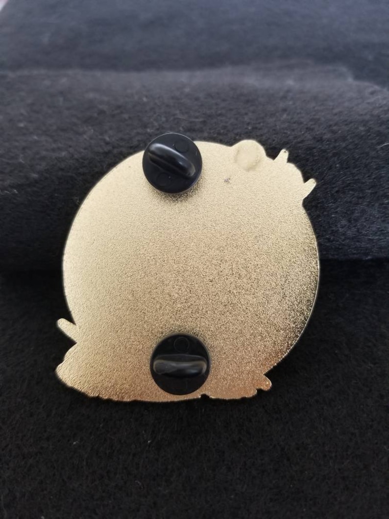 Puede incluir: La parte posterior de un pin de metal dorado con dos respaldos de goma negra. El pin presenta un dise&ntilde;o detallado de un personaje de dibujos animados.