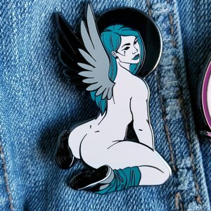 Puede incluir: Un pin de esmalte blanco y negro con un &aacute;ngel arrodillado con alas y cabello turquesa. El &aacute;ngel lleva un halo negro y tiene un cuerpo blanco.