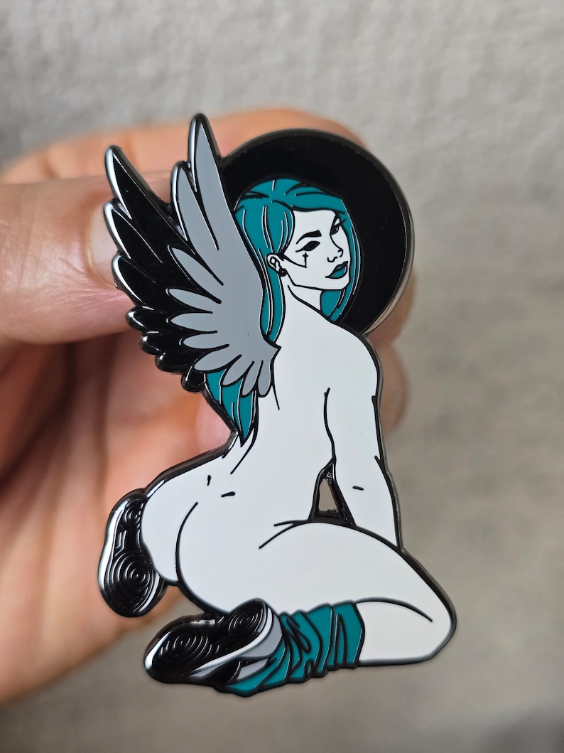 Puede incluir: Un pin de esmalte blanco y negro que representa a una mujer con cabello turquesa y alas. Est&aacute; arrodillada con una pierna doblada y la otra extendida. La mujer lleva un atuendo negro y turquesa. El pin tiene un fondo negro con un halo blanco.