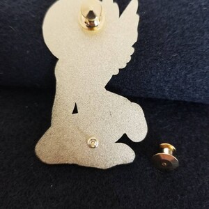 Puede incluir: Pin de metal dorado con un &aacute;ngel arrodillado con alas. El pin tiene un cierre de seguridad en la parte posterior.