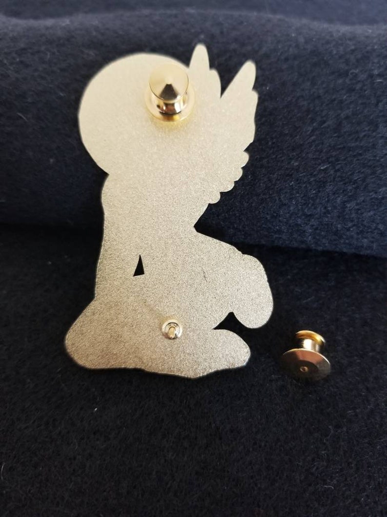 Puede incluir: Pin de metal dorado con un &aacute;ngel arrodillado con alas. El pin tiene un cierre de seguridad en la parte posterior.