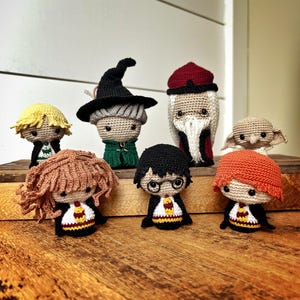 Op de afbeelding: Een verzameling van zeven handgemaakte gehaakte poppen, die personages uit een populaire fantasyserie uitbeelden. De poppen hebben gedetailleerde outfits en kapsels, waaronder een zwarte heksenhoed, rode gewaden en een ronde bril.