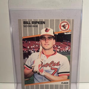 Könnte beinhalten: Baseballkarte mit Bill Ripken, Second Baseman der Baltimore Orioles. Die Karte zeigt Ripken in seiner Orioles-Uniform mit einem Schläger. Die Karte befindet sich in einer schützenden Plastikhülle. Der Text "Bill Ripken, Second Base" ist sichtbar.