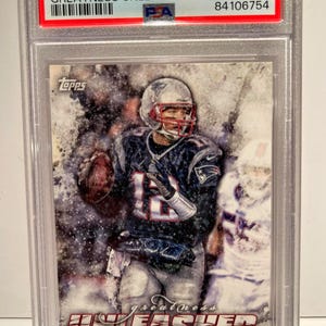 Könnte beinhalten: Eine Tom Brady Sammelkarte aus der 2014 Topps Greatness Unleashed Serie, bewertet mit GEM MT 10 von PSA. Die Karte zeigt ein Action-Foto von Brady in seinem Patriots-Trikot, mit dem Wort "UNLEASHED" prominent dargestellt.