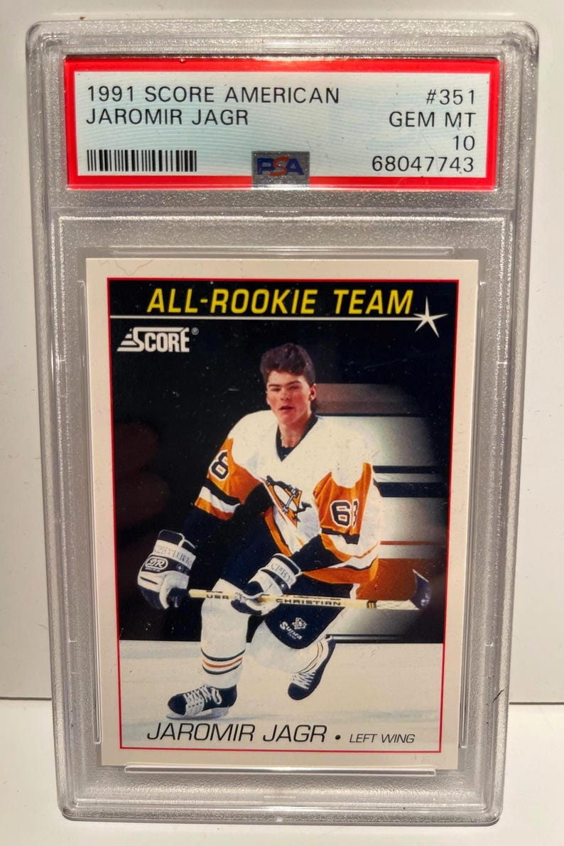 Puede incluir: Una tarjeta de hockey de Jaromir Jagr de 1991 Score American, calificada GEM MT 10 por PSA. La tarjeta presenta a Jagr con su uniforme de los Pittsburgh Penguins, con el texto "ALL-ROOKIE TEAM" y "SCORE". La tarjeta est&aacute; en un soporte protector.
