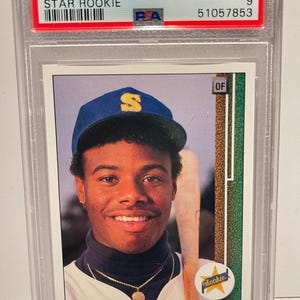 Puede incluir: Una tarjeta de béisbol de novato estrella de Ken Griffey Jr. de 1989 Upper Deck, calificada como #1 Mint 9 por PSA, en un soporte protector. La tarjeta presenta a Ken Griffey Jr. con una gorra azul.