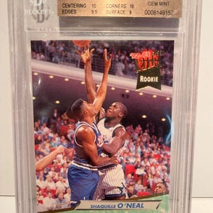 May include: A graded Shaquille O'Neal rookie card from the 1992-93 Ultra set, encased in a protective case. The card shows O'Neal in action, with the Orlando Magic logo. The card is graded 9.5 by Beckett.