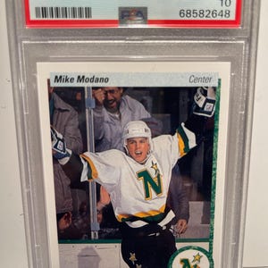 May include: A 1990 Upper Deck hockey card featuring Mike Modano of the Minnesota North Stars. The card is graded GEM MT 10 and has a PSA number of 68582648. The card shows Modano in his green and white uniform with the team's logo on the front. The card is in excellent condition.