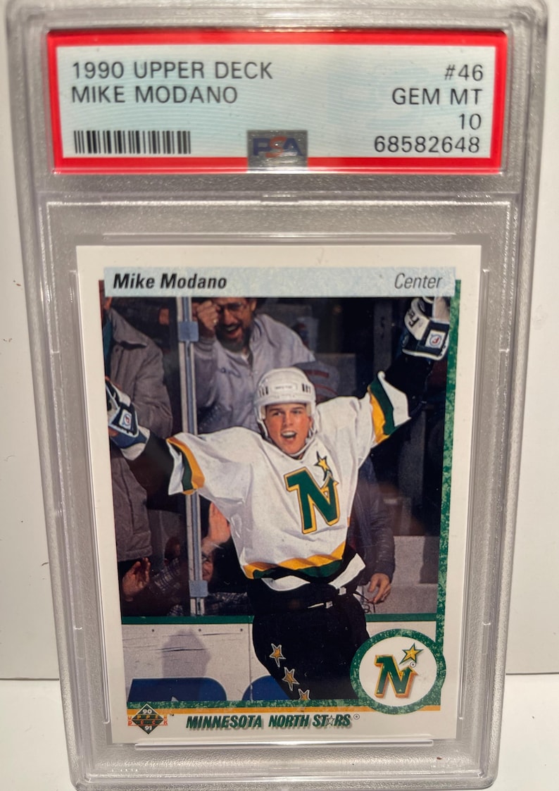 May include: A 1990 Upper Deck hockey card featuring Mike Modano of the Minnesota North Stars. The card is graded GEM MT 10 and has a PSA number of 68582648. The card shows Modano in his green and white uniform with the team's logo on the front. The card is in excellent condition.
