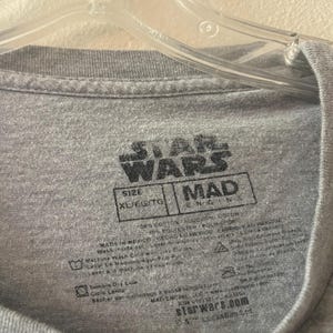 以下が含まれることがあります： グレーのスターウォーズTシャツタグで、黒いテキストが書かれています。タグには「スターウォーズ」と「マッドエンジン」とサイズ「XL/EG/TG」が書かれています。タグには、お手入れ方法とウェブサイト「starwars.com」も記載されています。