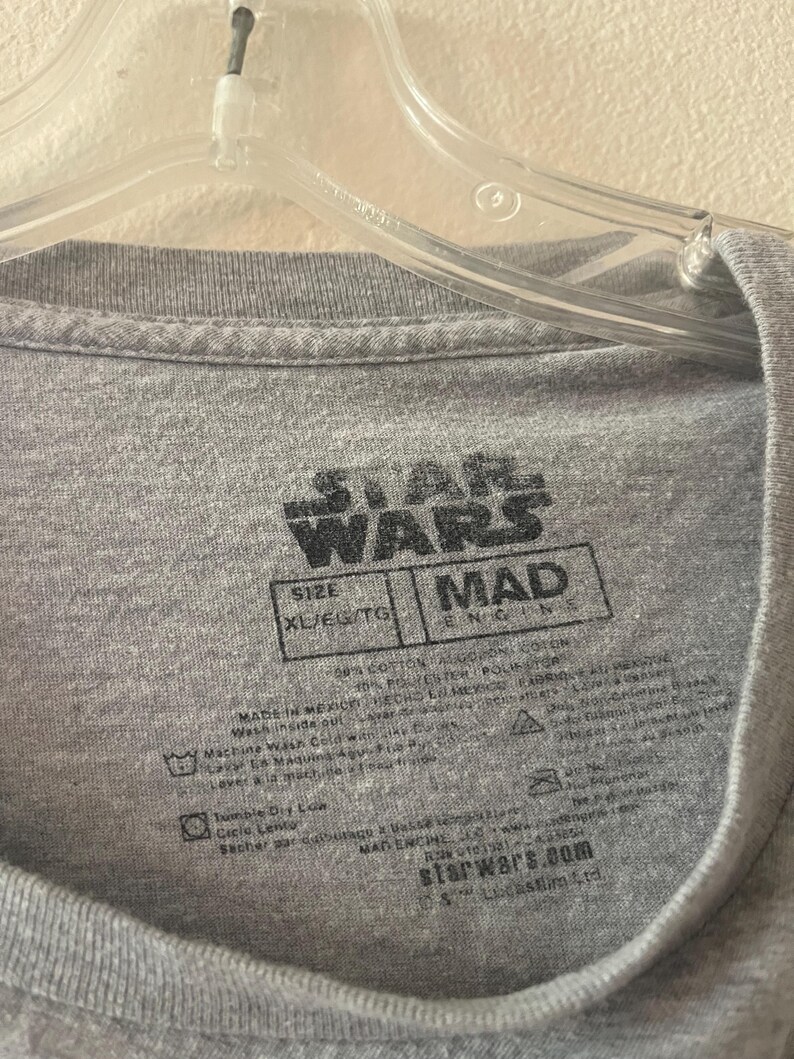 以下が含まれることがあります： グレーのスターウォーズTシャツタグで、黒いテキストが書かれています。タグには「スターウォーズ」と「マッドエンジン」とサイズ「XL/EG/TG」が書かれています。タグには、お手入れ方法とウェブサイト「starwars.com」も記載されています。