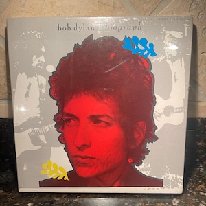May include: A 3-record deluxe edition vinyl record album cover featuring a red and white image of Bob Dylan with the text "bob dylan biograph" on the cover.