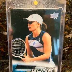 May include: A trading card featuring Iga Swiatek in a tennis pose, holding a racket. The card has a blue and white design with the text "Young Guns" and "Tennis" at the bottom. The card is encased in a clear protective holder.