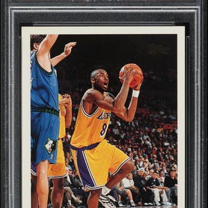 Könnte beinhalten: Eine 1996 Topps Kobe Bryant Basketballkarte, bewertet mit NM-MT 8 von PSA. Sie zeigt Kobe Bryant in einem gelb-violetten Lakers-Trikot in Aktion mit einem Basketball. Die Karte ist in einem Schutzhalter eingeschlossen.