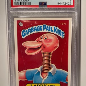 May include: A Garbage Pail Kids trading card featuring Larry Lips, a character with a pink head, a long, springy neck, and a wide, toothy smile. The card is red with white text and a blue border. The card number is 157a.