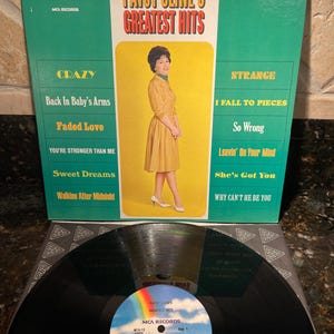 May include: A vintage vinyl record album titled "Patsy Cline's Greatest Hits." The album cover is green with a central image of Patsy Cline in a yellow dress. The record itself is black with a colorful label.