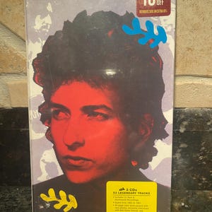 May include: A 3-CD set of Bob Dylan music featuring a yellow and blue design. The set includes 53 legendary tracks, 21 rare and unreleased recordings, and a 44-page colour book with photos, interviews, and track-by-track notes.
