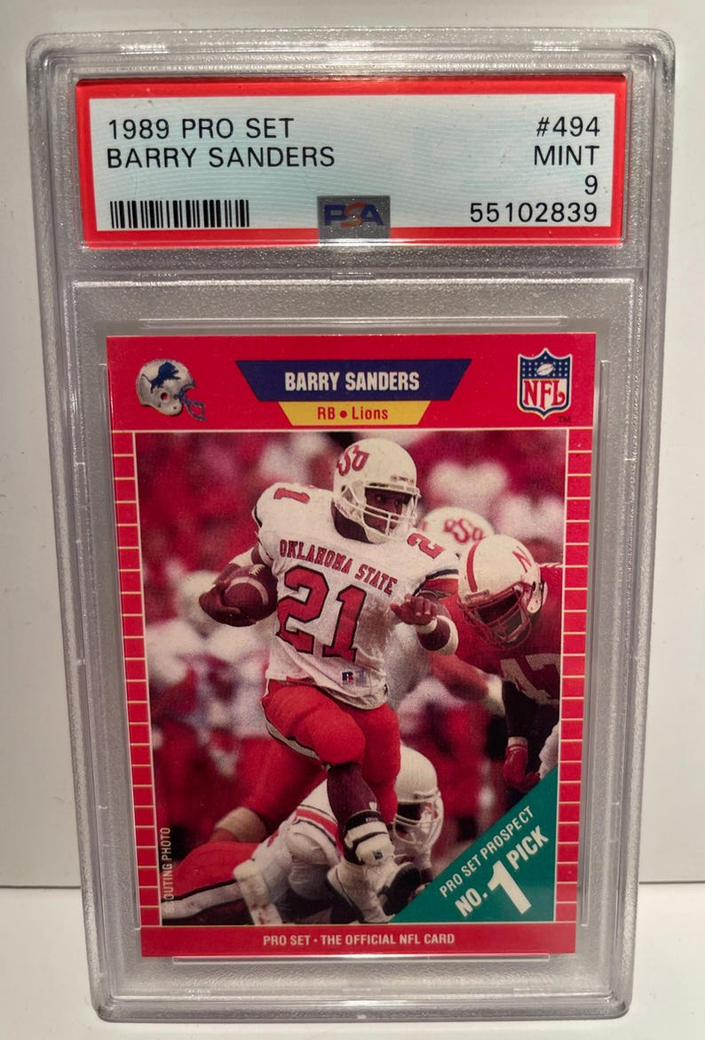 May include: A 1989 Pro Set Barry Sanders football card, graded as Mint 9. The card features a photo of Sanders in an Oklahoma State uniform, number 21, running with the ball. The card is encased in a protective holder.