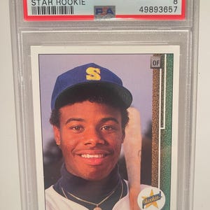 Puede incluir: Tarjeta de béisbol de novato estrella de Ken Griffey Jr. de 1989, Upper Deck, calificada NM-MT 8, en un soporte protector. La tarjeta muestra a Ken Griffey Jr. con una gorra azul con una "S" dorada y un bate de béisbol.