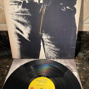 May include: The Rolling Stones' "Sticky Fingers" vinyl record album. The album cover features a black and white image of a pair of jeans with a working zipper. The record label is yellow with black text. The album is a classic rock music item.