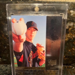 Puede incluir: Una tarjeta de béisbol con un jugador de los Giants sosteniendo una pelota de béisbol y usando una gorra. La tarjeta está dentro de un protector transparente. Las palabras "ROOKIE CARD" y "GIANTS" son visibles en la tarjeta.