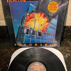 Könnte beinhalten: Def Leppard's Vinyl-Schallplatte "Pyromania". Das Albumcover zeigt ein Fadenkreuz über einer Stadtlandschaft mit einer feurigen Explosion. Die Schallplatte ist schwarz mit einer weißen Papierhülle.