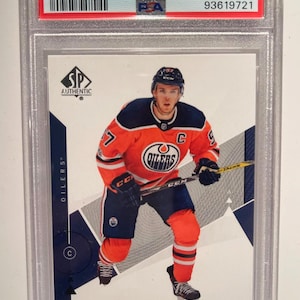 May include: A 2018 SP Authentic Connor McDavid hockey card, graded GEM MT 10. The card shows McDavid in an Edmonton Oilers jersey, holding a hockey stick. The card is encased in a protective holder.