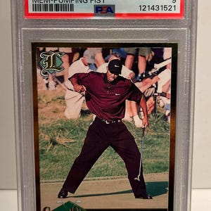 Puede incluir: Una tarjeta de intercambio deportiva calificada con Tiger Woods. La tarjeta muestra a Woods con una camisa burdeos y pantalones negros, a mitad de swing. La tarjeta est&aacute; etiquetada como "2001 Legends Sports" y "Mem-Pumping Fist."