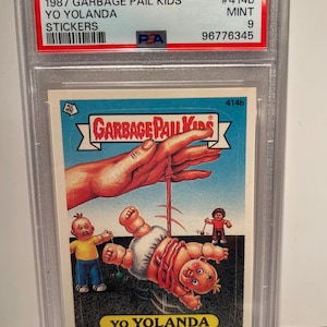 May include: A graded Garbage Pail Kids trading card featuring a cartoon baby doll with red hair and a stitched-up face. The baby doll is hanging upside down from a string held by a giant hand. The card is graded 9 by PSA and is from the 1987 series. The card is titled "Yo Yolanda" and has the number 414b.