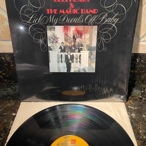 May include: A black vinyl record album cover with a red and white design. The album is titled "Lick My Decals Off, Baby" by Captain Beefheart & The Magic Band. The cover features a photo of the band in a grand setting.