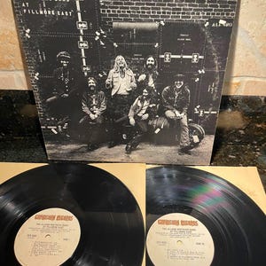 May include: A black and white album cover for The Allman Brothers Band at Fillmore East, featuring a band photo. Two vinyl records are visible below, with labels reading "Capricorn Records" and the album title.