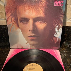 May include: A vinyl record album cover featuring a close-up portrait of David Bowie. The album is titled "Space Oddity" and features the RCA Victor label. The album cover is a light brown color with a pink background.