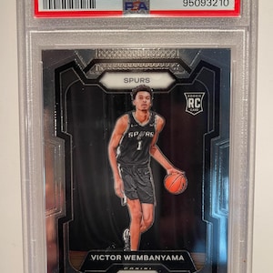 May include: A 2023 Prizm Victor Wembanyama rookie card, graded Gem Mint 10, encased in a protective holder. The card features a player in a Spurs uniform with a basketball. The card's details include the player's name and the Prizm logo.