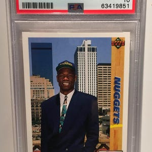 Può includere: Una carta da basket Dikembe Mutombo Upper Deck del 1991, valutata GEM MT 10 da PSA. La carta presenta un ritratto di Mutombo in uniforme Nuggets, con il logo della squadra e il logo NBA Draft.