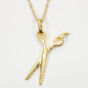May include: A gold-colored necklace featuring a scissors pendant. The scissors are detailed with curved handles and sharp blades, suspended from a delicate chain. The pendant is a charm, likely for a hairstylist or barber.