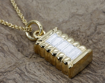 Puede incluir: Un collar de oro con un colgante con forma de pequeño edificio. El colgante está hecho de oro y plata y tiene una superficie texturizada.