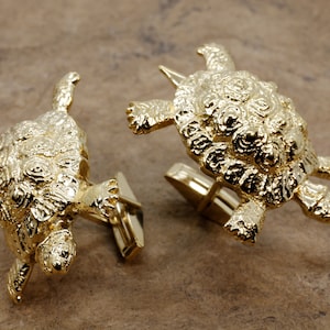 May include: A pair of gold-colored cufflinks, each shaped like a detailed turtle. The turtles have textured shells and outstretched limbs. The cufflinks are displayed on a brown surface.