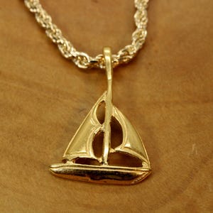 Può includere: Un ciondolo in metallo dorato a forma di barca a vela con due vele distinte e uno scafo, sospeso a una catena a corda abbinata color oro. Il ciondolo presenta una superficie lucida e riflettente che cattura la luce, mettendo in risalto il design nautico.