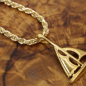 Può includere: Una collana color oro con catena a corda e un ciondolo a forma di barca a vela. Il ciondolo ha una finitura lucida con sezioni ritagliate che rappresentano le vele e lo scafo, sospeso tramite un occhiello situato sulla cima dell'albero.