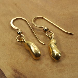 May include: A pair of gold-colored dangle earrings. Each earring features a curved hook, a small ball, and a bean-shaped charm. The earrings are displayed against a light brown background.