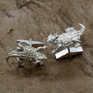 May include: A pair of silver cufflinks featuring a detailed, textured design of a horned lizard. The cufflinks are a unique and stylish accessory for any occasion.