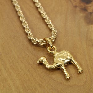 May include: A gold-colored necklace featuring a camel pendant. The pendant is a detailed, three-dimensional camel figure. The chain is a thick, rope-style design. The necklace is displayed on a wooden surface.