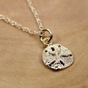 Peut inclure: Un collier en or avec un petit pendentif en forme de dollar des sables. Le pendentif a une surface texturée et une étoile à cinq branches au centre.